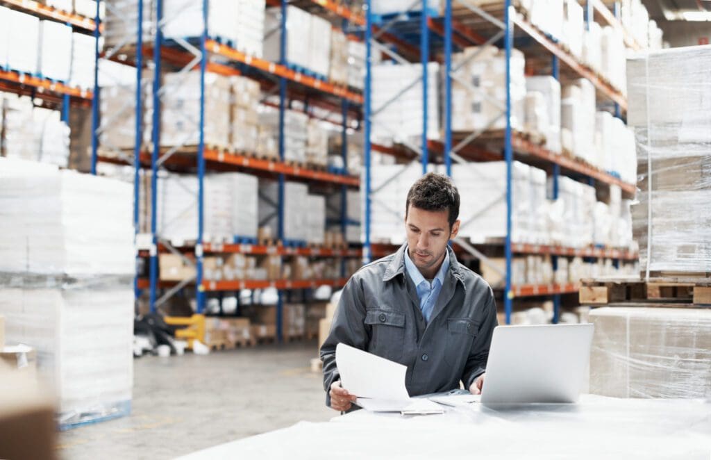 A young man using his laptop and looking over his notes while working in a warehouse